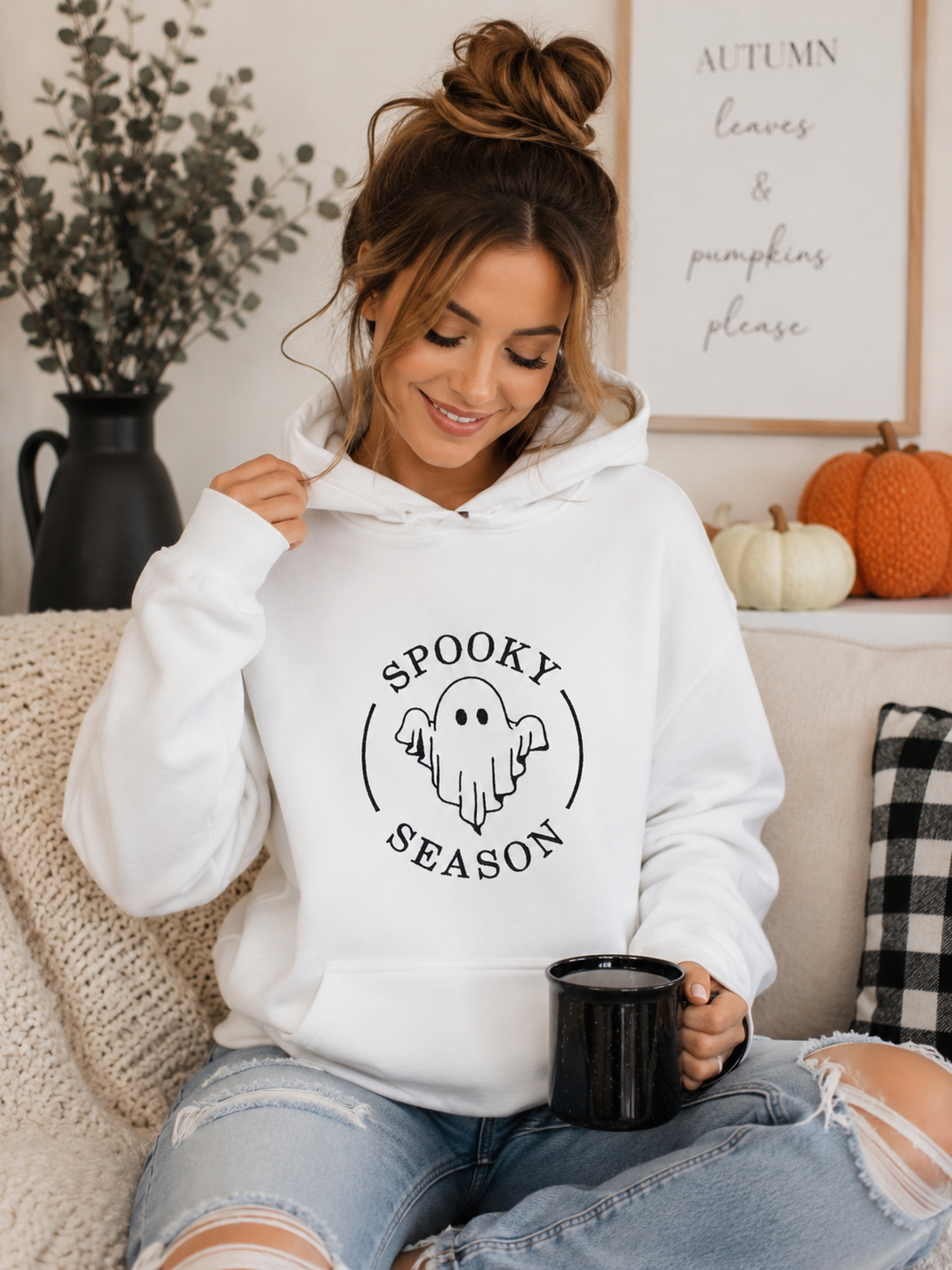 Model wearing white hoodie with Spooky Season ghost embroidery on chest, sitting on a couch with fall decor