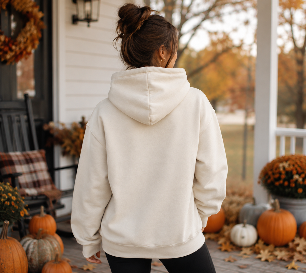 A woman viewed from behind wearing a cream-colored hoodie on a porch decorated for autumn, with pumpkins, fall leaves, and a rocking chair in the background.
