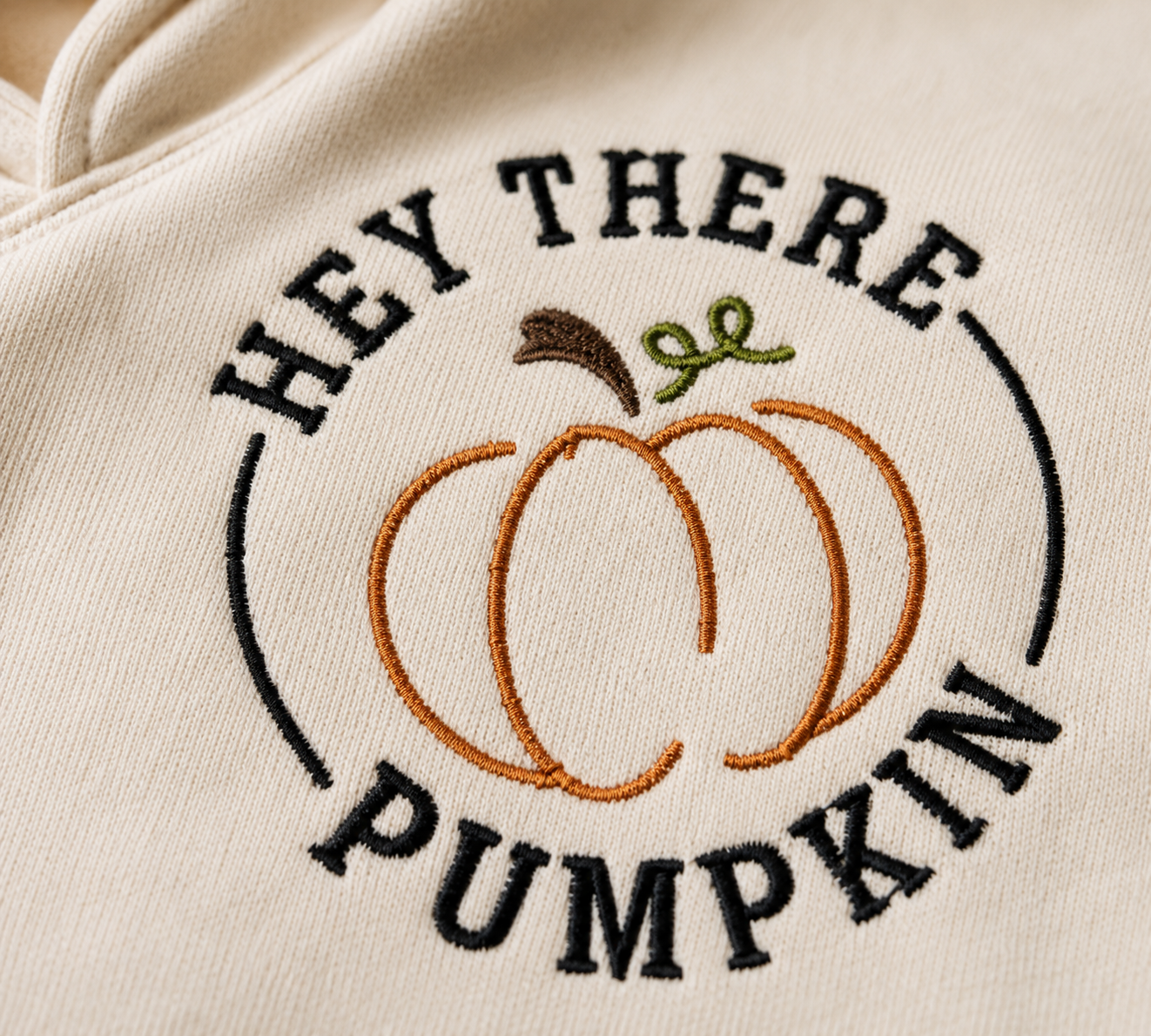 Close-up of a cream-colored hoodie featuring an embroidered design of a pumpkin with the text 'HEY THERE PUMPKIN' in black, orange, brown, and green.