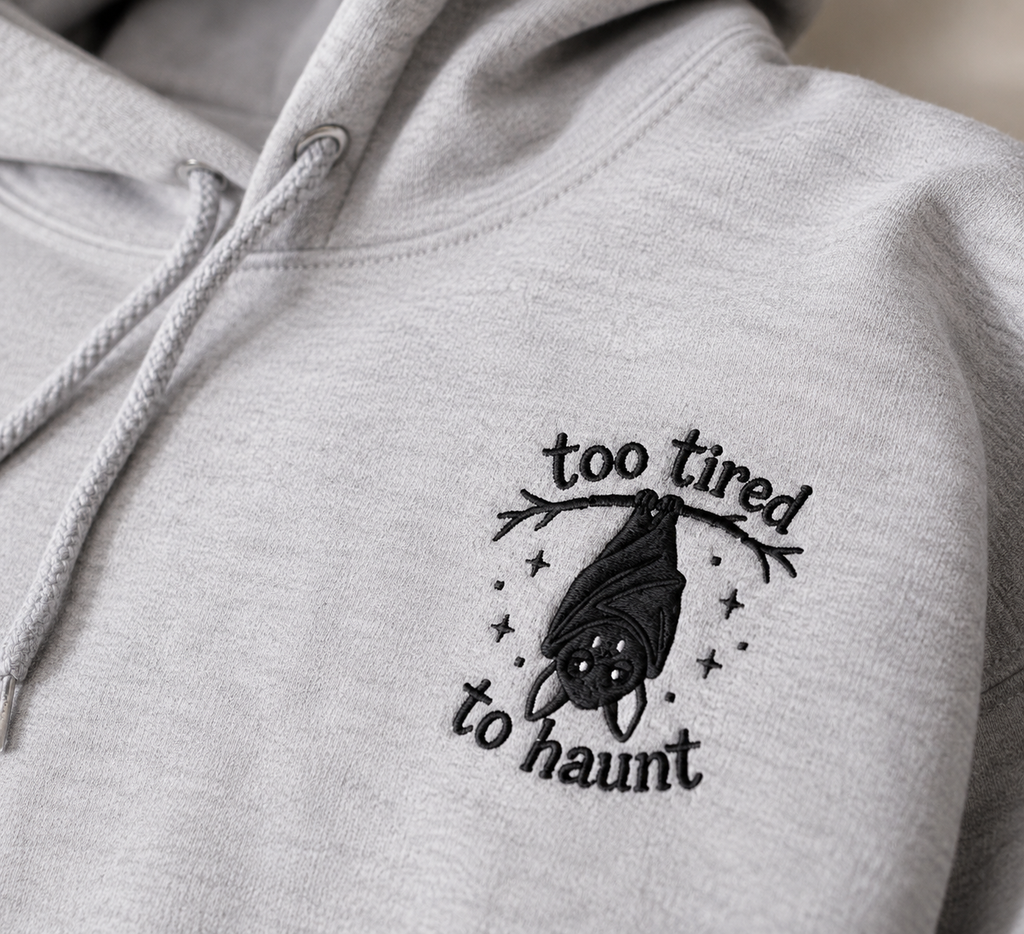 Close-up of heather gray hoodie with 'too tired to haunt' bat embroidery on left chest