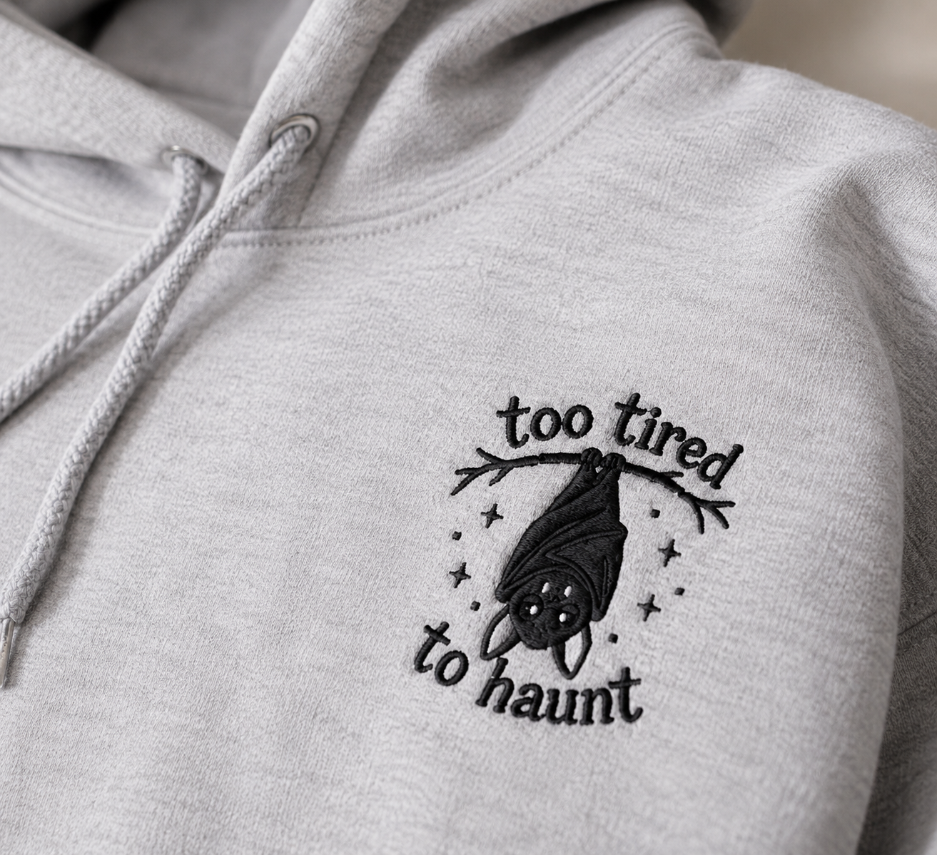 Close-up of heather gray hoodie with 'too tired to haunt' bat embroidery on left chest