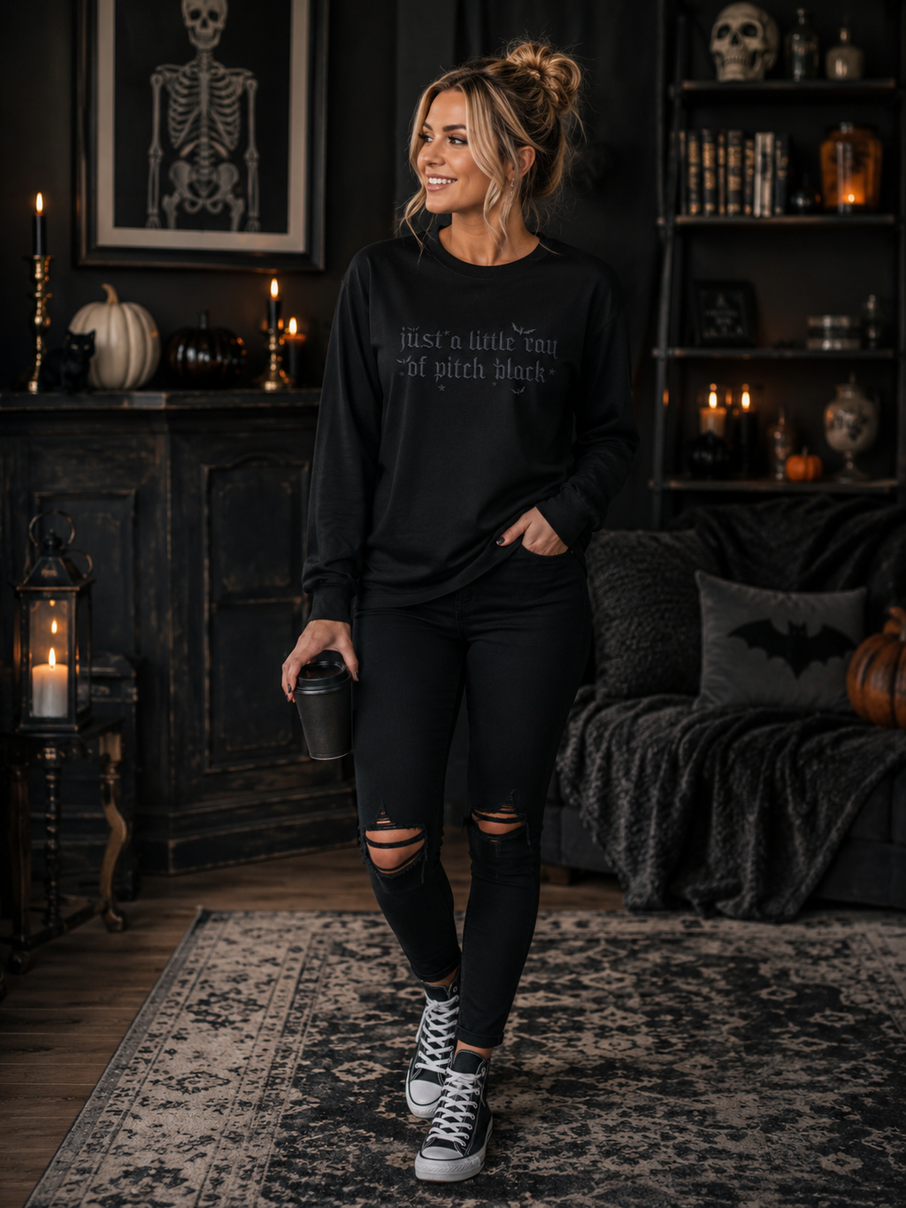 A woman stands in a dark, gothic-themed room wearing a black long-sleeve crewneck sweatshirt with the text 'just a little tiny bit of pitch black' printed in white on the chest, paired with black ripped leggings and black high-top sneakers.