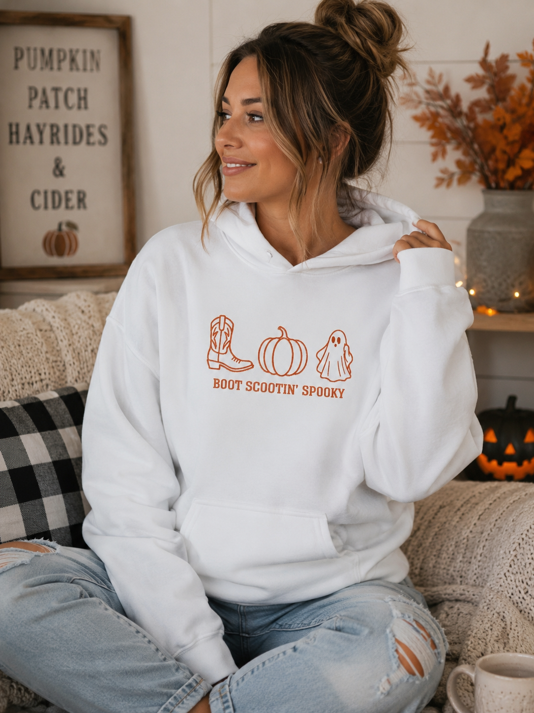 A woman wearing a white pullover hoodie with a pumpkin, cowboy boot, and ghost graphic in orange, sitting on a cozy couch in a fall-themed room.