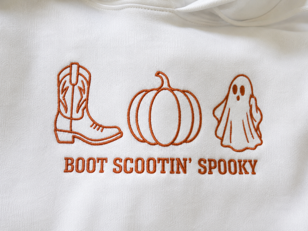 Close-up of a white garment with orange embroidered icons of a cowboy boot, pumpkin, and ghost above the text 'BOOT SCOOTIN' SPOOKY'.