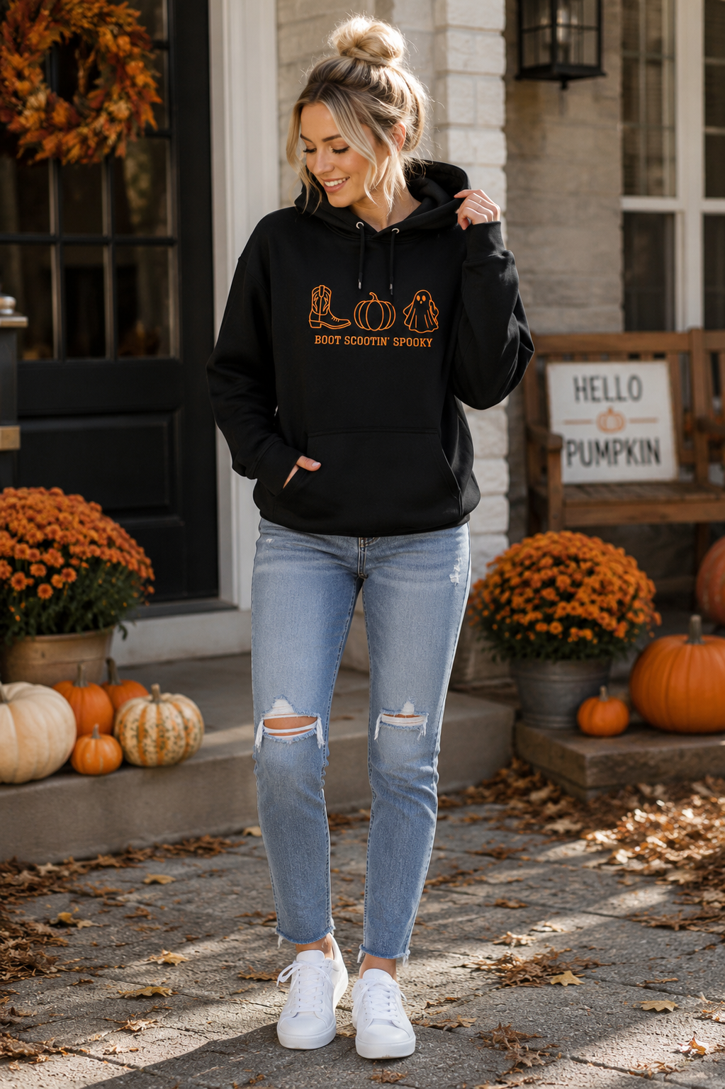A woman stands on a front porch wearing a black pullover hoodie with a gold graphic that reads 'MOST FAVORITED SPOOKTIFTER' beneath pump and pumpkin illustrations. She is also wearing light-wash ripped jeans and white sneakers.
