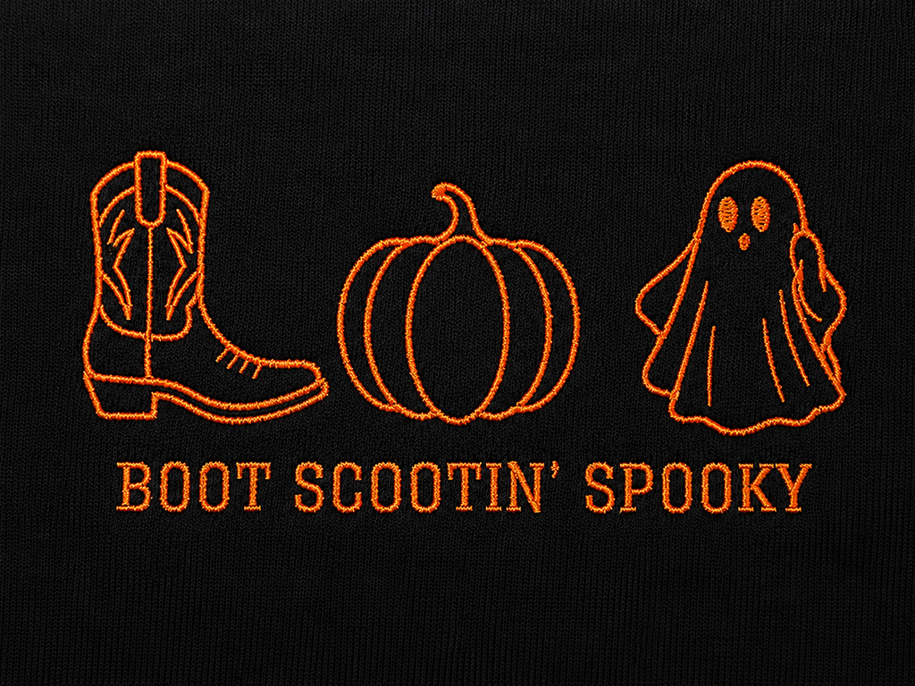 Close-up of embroidered orange Halloween-themed graphics on black fabric, including a cowboy boot, pumpkin, ghost, and the text 'BOOT SCOOTIN' SPOOKY'.