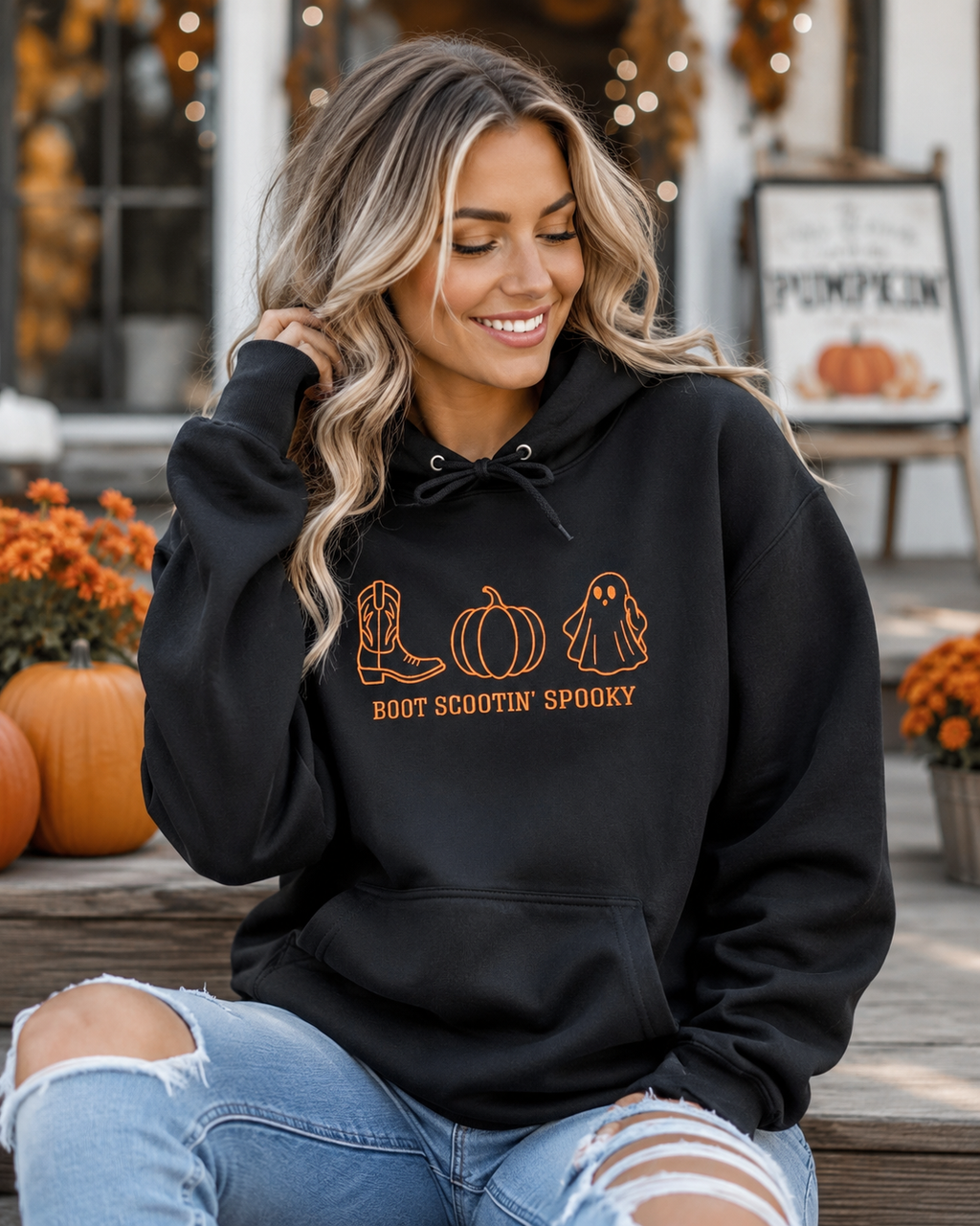 A woman wearing a black hoodie with a 'Boot Scootin' Spooky' graphic sits on a wooden bench in front of a storefront decorated with pumpkins and fall lights.