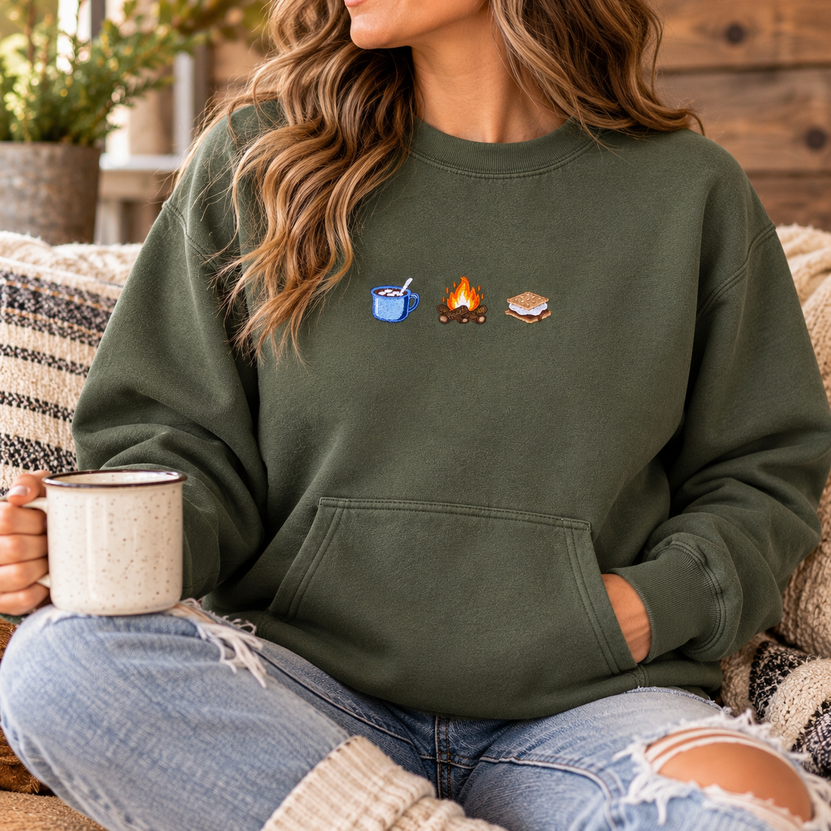 Seated model wearing an olive-green crewneck with mug, campfire, and s’more embroidery and a kangaroo pocket.