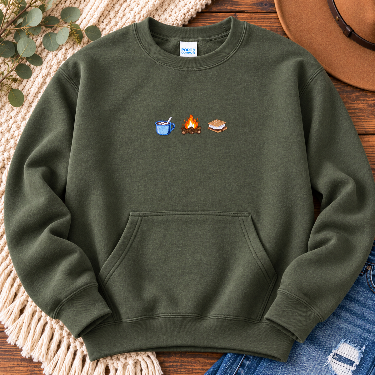 Olive-green embroidered kangaroo-pocket crewneck laid flat on wood beside a knit blanket, brown hat, and jeans.